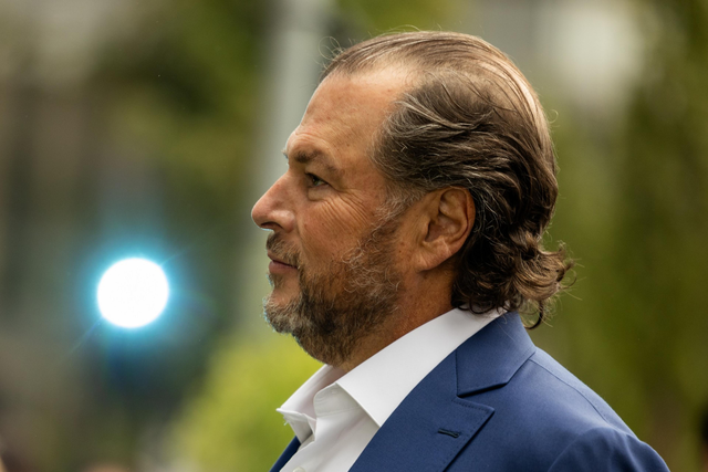 A man with slicked-back hair and a beard wears a blue suit and white shirt, facing left with a soft green and light background behind him.